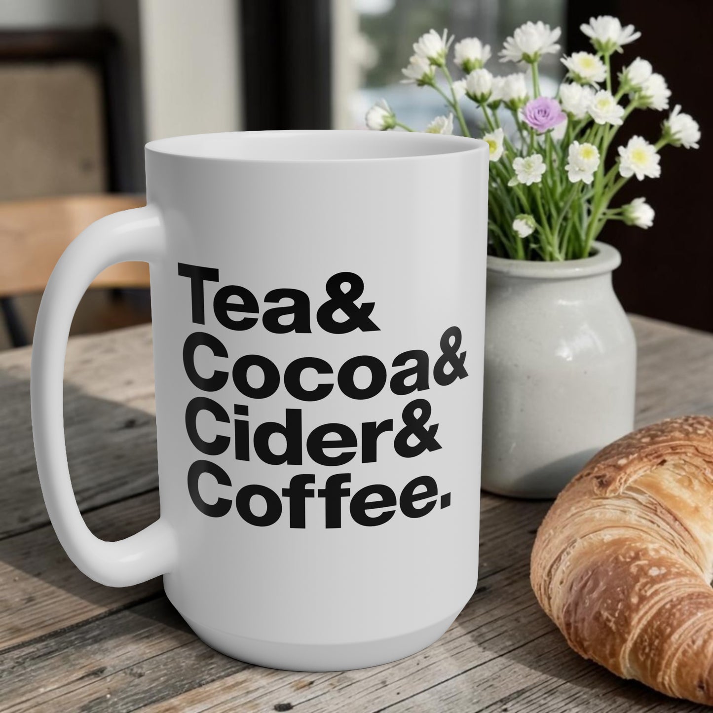White mug with text 'Tea & Cocoa & Cider & Coffee' on a wooden table with flowers and bread.