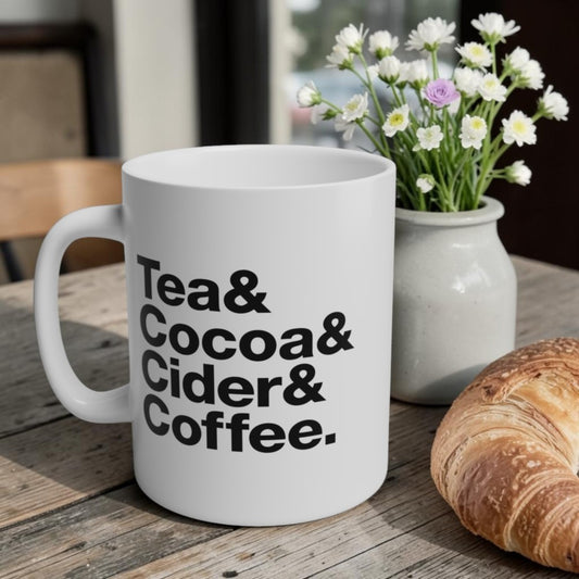 White mug with text 'Tea & Cocoa & Cider & Coffee' on a wooden table with flowers and bread.